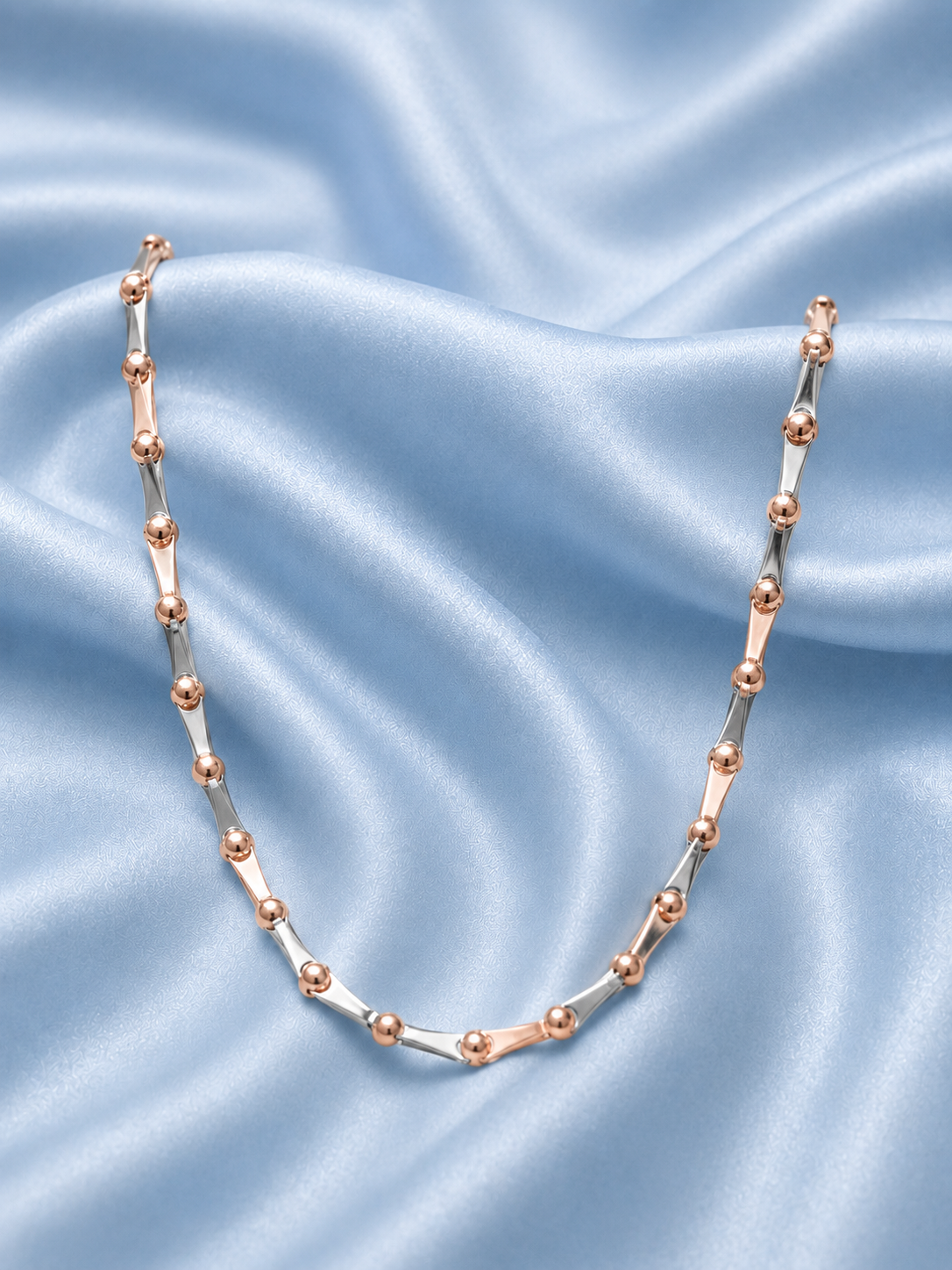 A delicate necklace with alternating polished rose gold and silver cylindrical beads, arranged in a U-shape on a draped light blue satin fabric background.