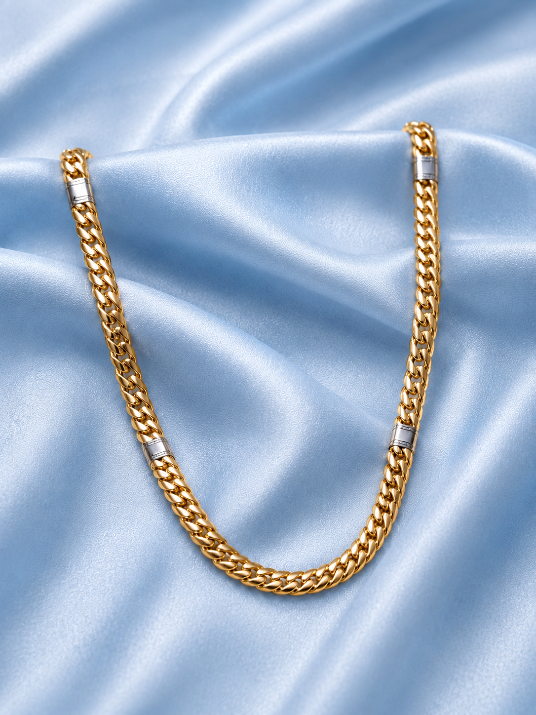 A gold Cuban link chain necklace with silver-toned accent rings, arranged in a U-shape on a light blue satin fabric background.