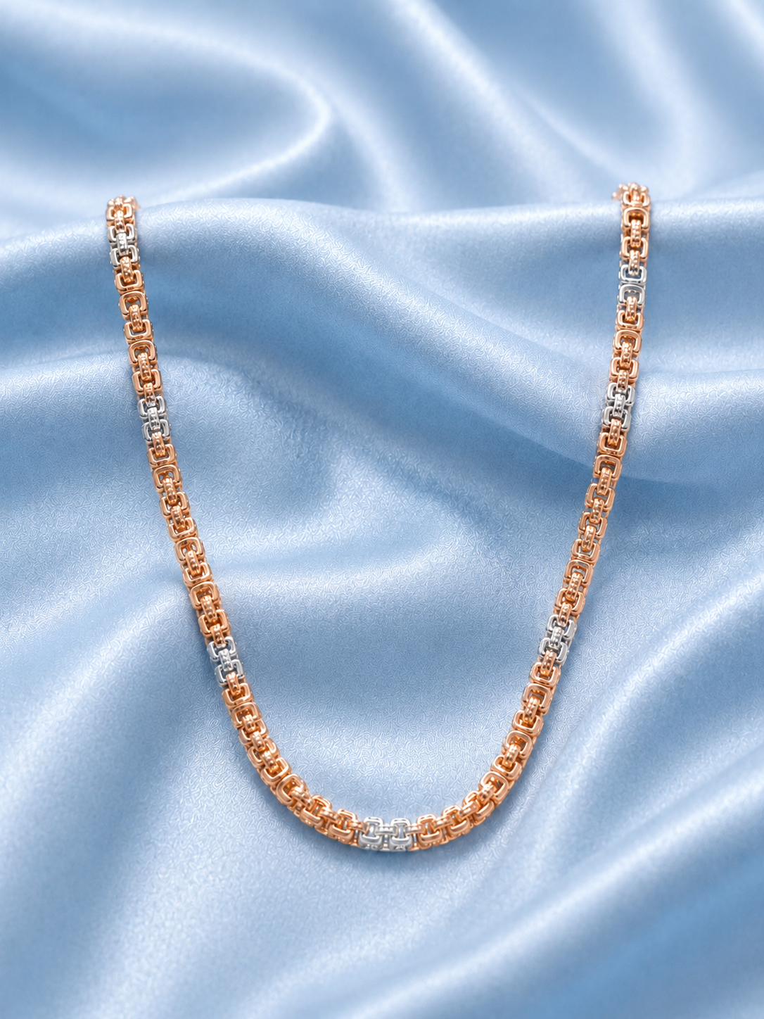 A tennis-style chain necklace with alternating rose gold and silver-toned links, displayed on a draped light blue satin fabric background.