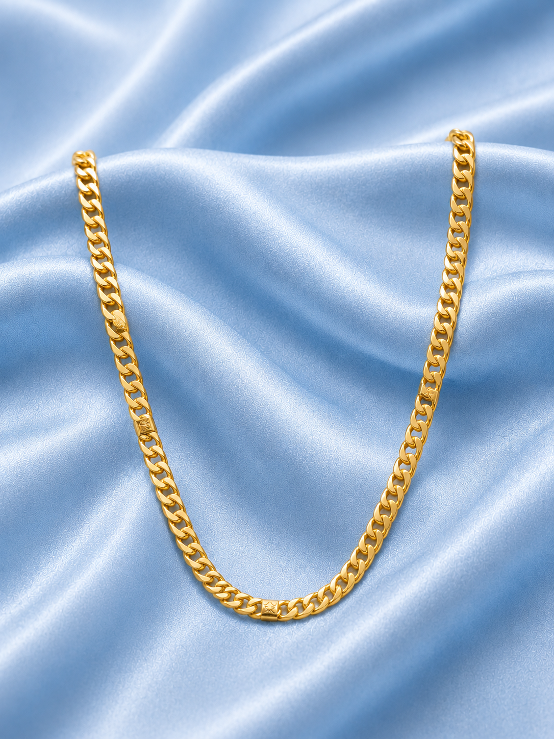 A gold Cuban link chain necklace draped in a U-shape over light blue satin fabric.
