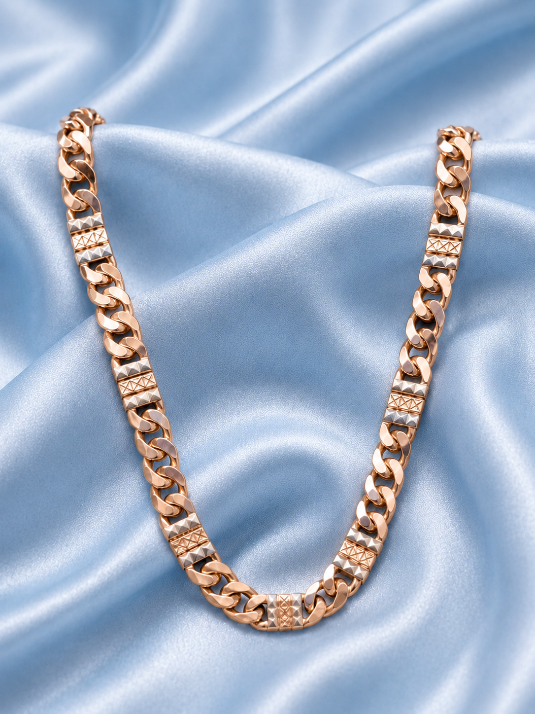 A rose gold Cuban link chain necklace with textured and diamond-like accent links, displayed on a draped light blue satin fabric background.