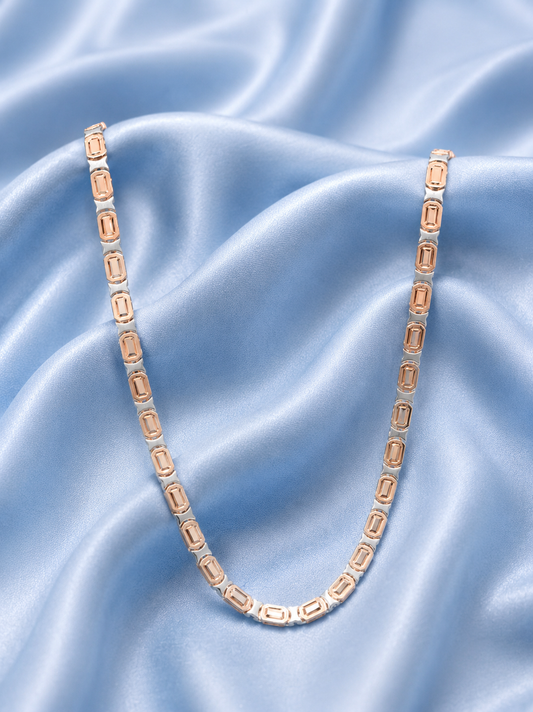 A delicate tennis-style chain necklace with elongated rectangular links, featuring a warm rose-gold-toned center and silver-toned outer edges, displayed on draped light blue satin fabric.