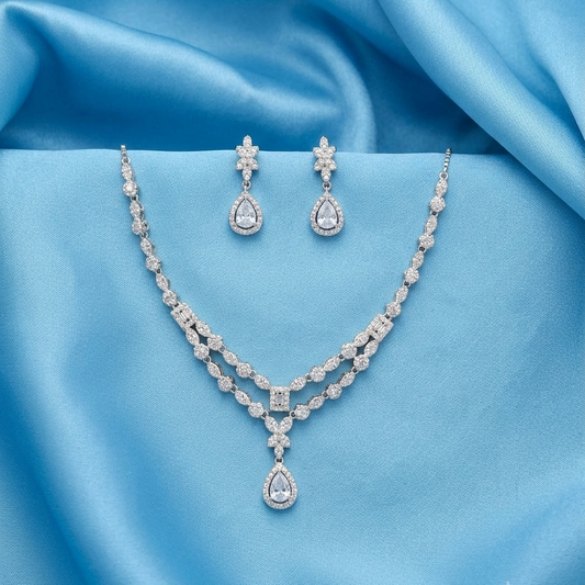A silver-tone jewelry set featuring a double-strand necklace and matching drop earrings, both adorned with clear stones, displayed on a draped light blue satin fabric background.
