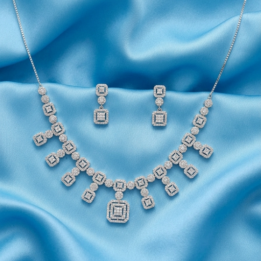A silver-toned jewelry set featuring a necklace and matching drop earrings, both adorned with clear gemstones in a geometric square pattern, displayed on a draped light blue satin fabric background.