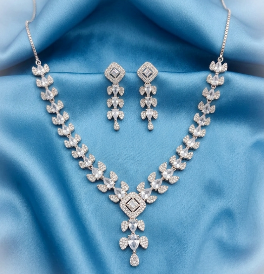 A silver-tone jewelry set featuring a necklace and matching earrings, both adorned with clear crystals, displayed on a draped light blue satin fabric background.