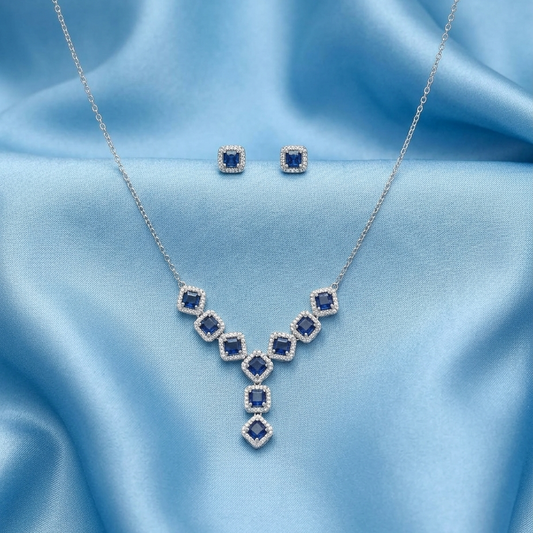Silver necklace and matching stud earrings with blue square-cut stones, displayed on a light blue satin fabric background.