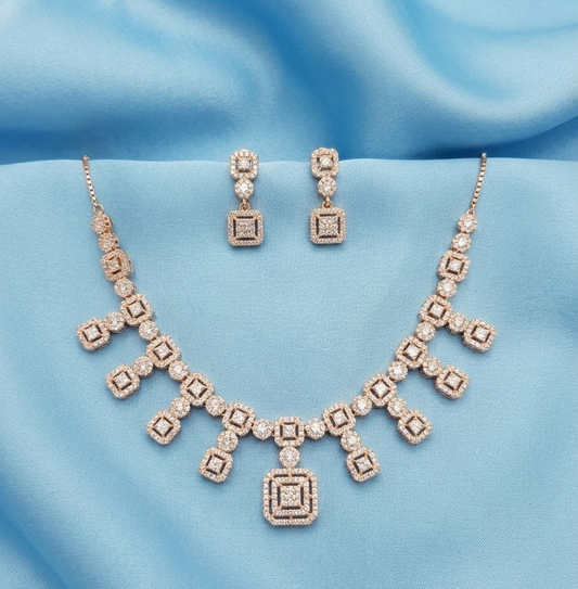 A gold-plated jewelry set featuring a necklace and matching drop earrings, both adorned with square-cut clear gemstones, displayed on a draped light blue satin fabric.