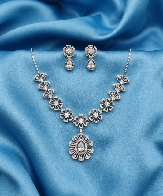 A silver-toned traditional Indian jewelry set featuring a necklace and matching earrings, decorated with clear teardrop and round stones, displayed on a draped blue satin fabric background.