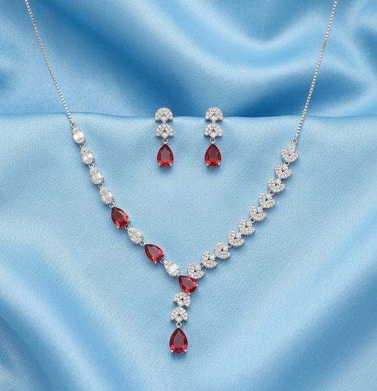 Silver-tone necklace and matching drop earrings set with clear crystals and red teardrop gemstones, displayed on a light blue satin fabric background.