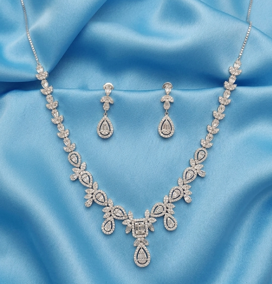 A silver-toned jewelry set featuring a necklace, matching earrings, and a pendant, all adorned with clear stones and arranged on a draped light blue satin fabric background.